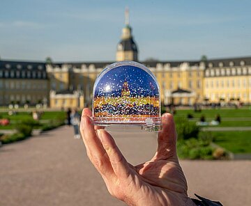 Eine Schneekugel mit einem Bild des Karlsruher Schlosses wird in das Bild gehalten. Im Hintergund ist unscharf das echte Schloss zu sehen.