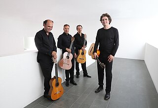 Vier Personen mit Gitarren stehen in einem weißen Raum.