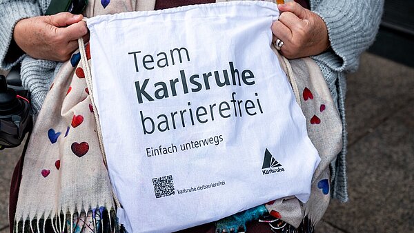 Person im Rollstuhl hält einen Stoffbeutel mit der Aufschrift "Team Karlsruhe barrierefrei"