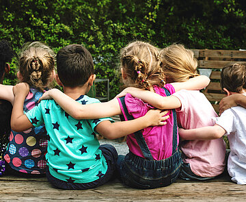 Bunt gekleidete Kinder im Kindergartenalter von hinten, legen sich gegenseitig die Arme um die Schultern