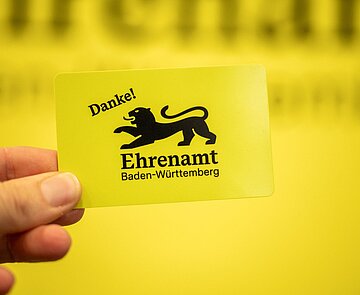 Hand hält gelbe Ehrenamtskarte Baden-Württemberg 