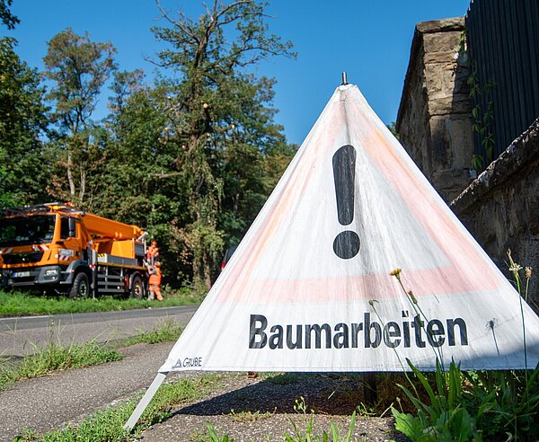 Nahaufnahme des Schilds "Baumarbeiten". Im Hintergrund sind die Baumpfleger-/innen auf dem Hubsteiger zu sehen.