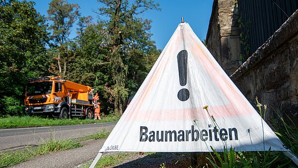 Nahaufnahme des Schilds "Baumarbeiten". Im Hintergrund sind die Baumpfleger-/innen auf dem Hubsteiger zu sehen.