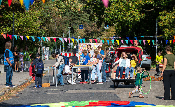 Karlsruhe spielt 2026: Blick auf eine Straße mit spielenden Kindern, seitlich Bürgermeisterin Melchien mit einem Mädchen in einer Rikscha.
