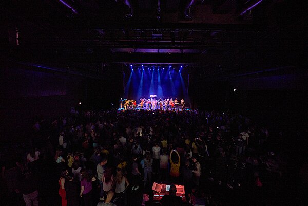 Foto vom Finale des Musikwettbewerbs 'Ecole du Flow' während der Europawochen, aufgenommen im Tollhaus. Das Bild zeigt eine belebte Szene mit vielen Zuschauern, die einer Performance auf einer beleuchteten Bühne in einer dunklen Halle beiwohnen. 