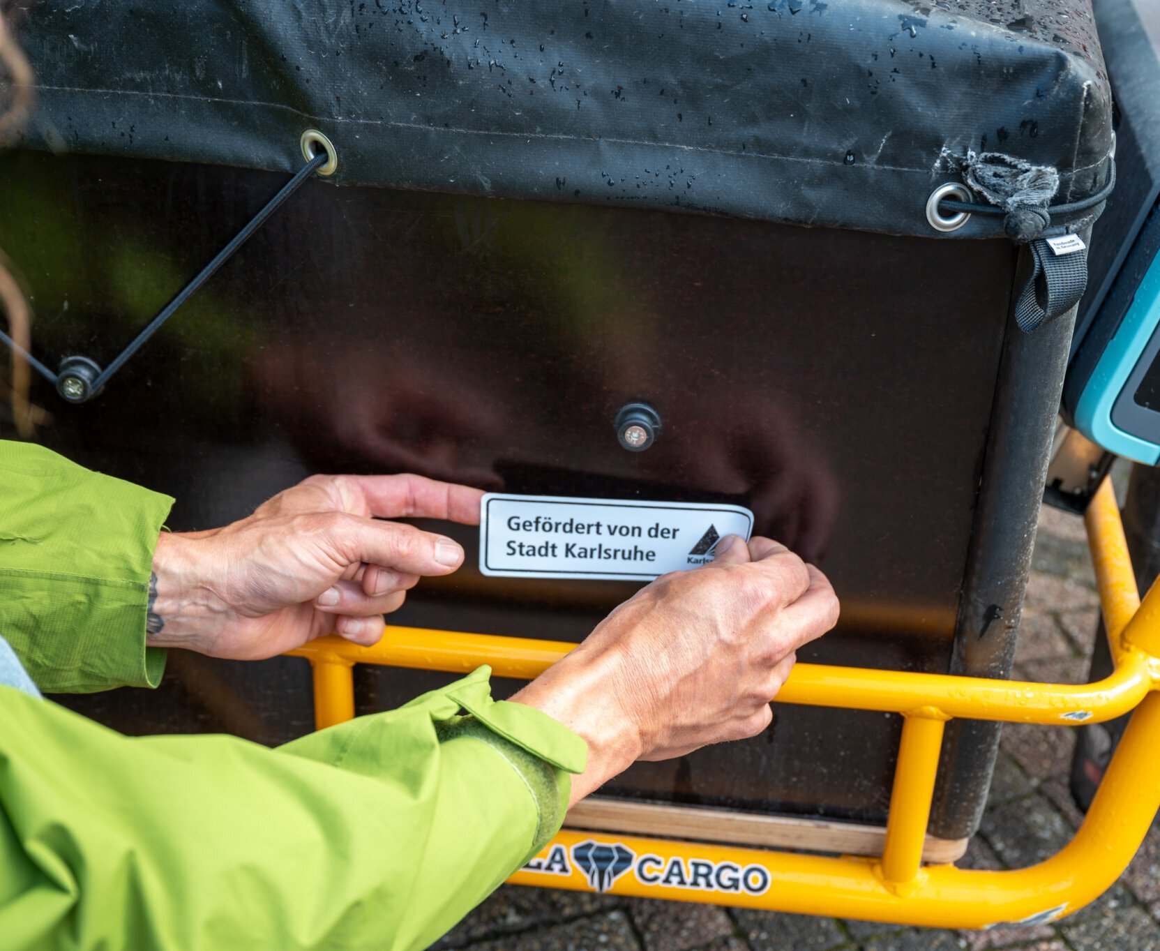 ein Aufkleber " gefördert von der Stadt Karlsruhe" wird auf Lastenrad geklebt