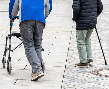 Zwei ältere Menschen mit Gehbehinderung laufen mit Rollator und Stock über Marktplatz