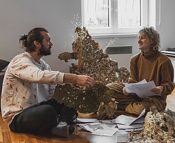 Ein Mann und eine Frau auf dem Boden sitzend umgeben von vielen bedruckten Papieren und kartonartigen Gebilden