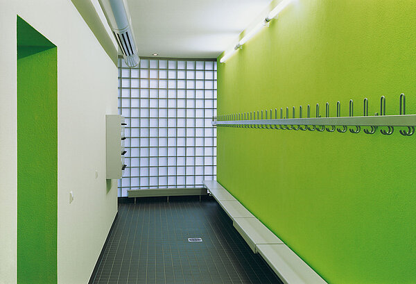 Zu sehen ist eine Umkleidekabine in auffälligem grün-weißen Anstrich, die mit einer Garderobe und Sitzbänken ausgestattet ist.