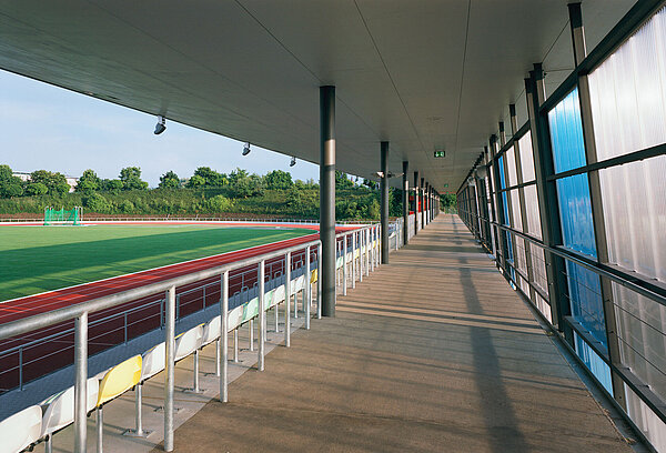 Blick entlang des überdachten oberen Tribünenwegs mit verglaster westlicher Rückwand und auf der gegenüberliegenden Seite abgrenzendem Geländer zu den Sitzschalen.