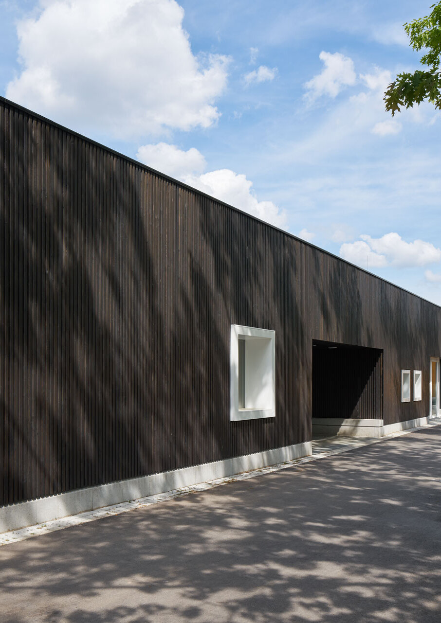 Zu sehen ist die Südwestansicht der grau lasierten Holzlattenfassade mit einem hellgrauen Sockelstein und mehreren Fenstern mit kräftigen weißen Holzrahmen.