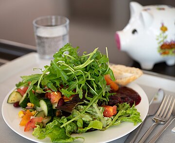 Teller mit  Salat auf Tablett und Sparschwein im Hintergrund in der Karlskantine