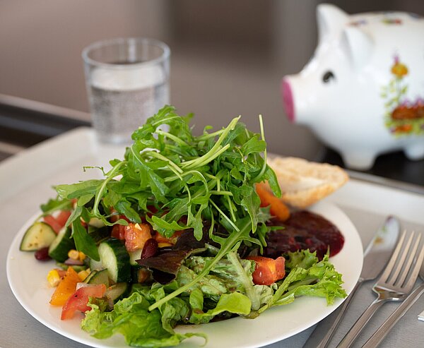 Teller mit  Salat auf Tablett und Sparschwein im Hintergrund in der Karlskantine