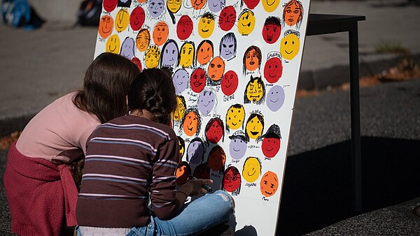 Zwei Kinder sitzen vor einer selbstgestalteten Wand mit Gesichtern  im Rahmen der Aktion Karlsruhe spielt! 2026 und zeichnen