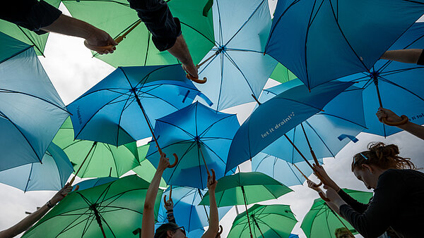Das Bild zeigt viele blaue und grüne Regenschirme, die im Rahmen einer spontanen Aktion, so genannter FreshMob in die Luft gehalten werden. Diese sollen schattenspendende Bäume symbolisieren, die in der Innenstadt, hier am Beispiel der Herrenstraße, mit Blick auf den Klimawandel benötigt werden.