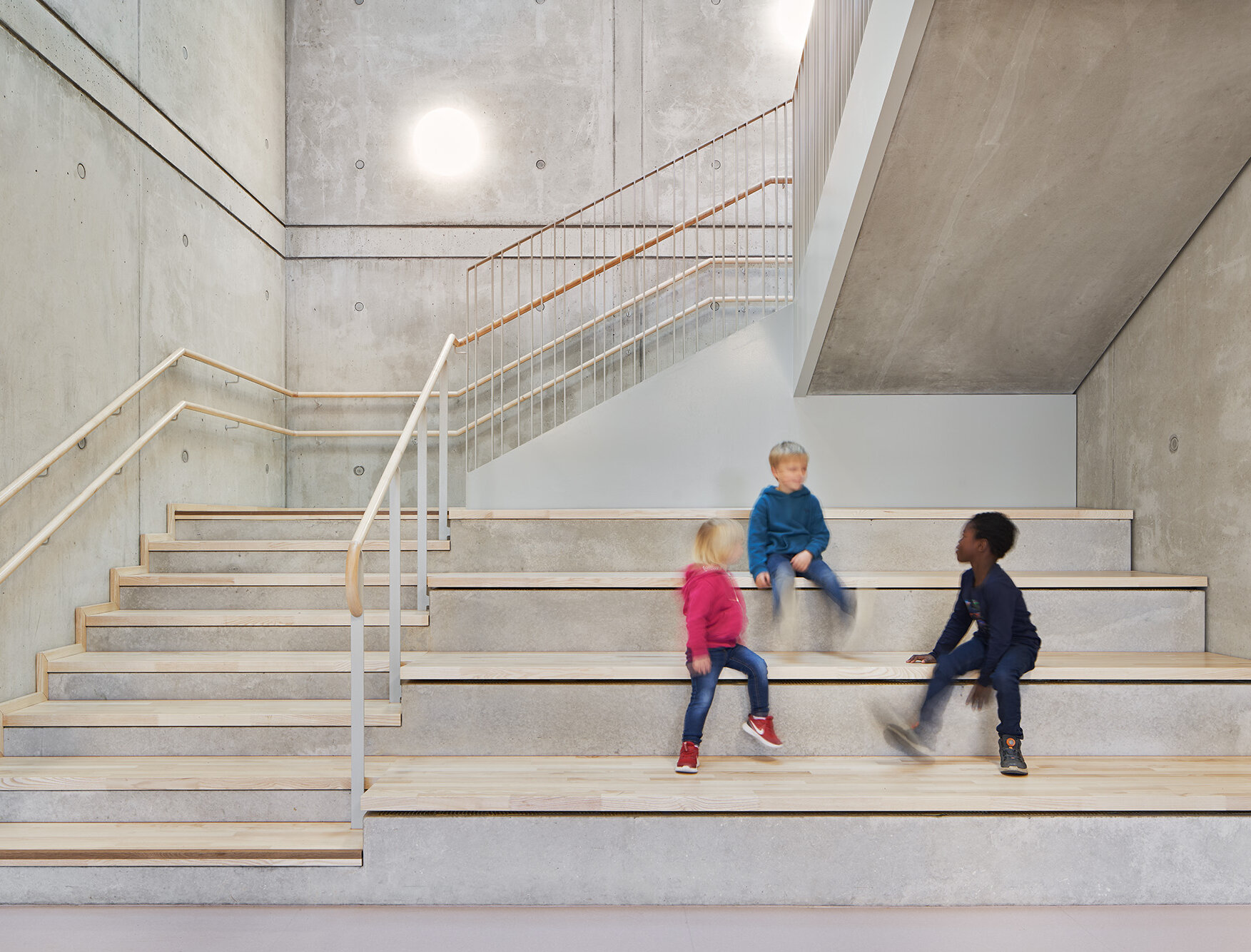 Das Foto zeigt Sichtbetonwände, eine Treppe mit Holzbelag und -handlauf sowie Sitzstufen, auf denen Kinder sitzen.