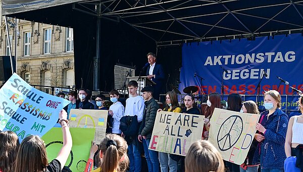 Schülerinnen und Schüler stehen beim Aktionstag gegen Rassismus mit Plakaten vor einer Bühne, auf der Oberbürgermeister Dr. Frank Mentrup spricht.