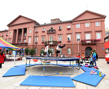 Vor dem Rathaus steht in Trampolin. Darauf springen Kinder.
