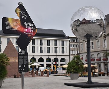 Ein Hinweispfeil auf dem Marktplatz mit der Aufschrift "Medienkunst ist hier" zeigt auf eine Discokugel.