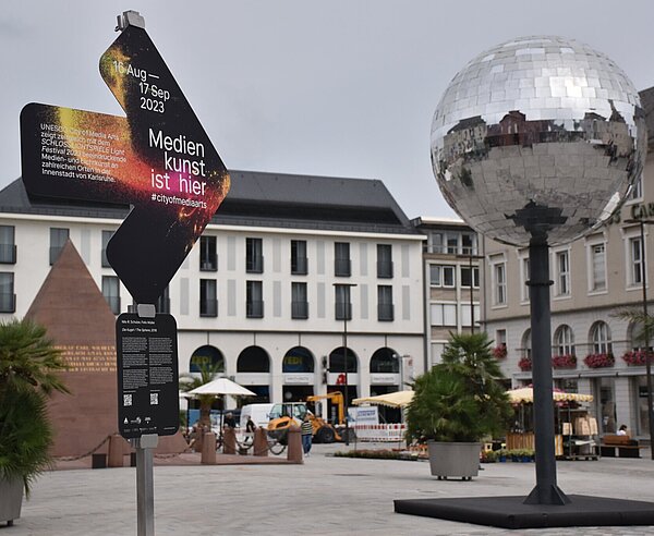 Ein Hinweispfeil auf dem Marktplatz mit der Aufschrift "Medienkunst ist hier" zeigt auf eine Discokugel.