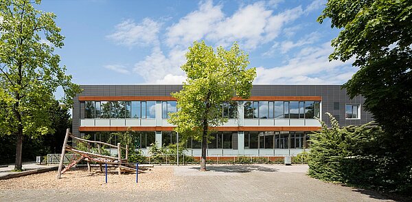Das Bild zeigt den Schulhof und die anthrazitfarbene Südfassade, die mittig von einem kleinen Baum bedeckt ist.