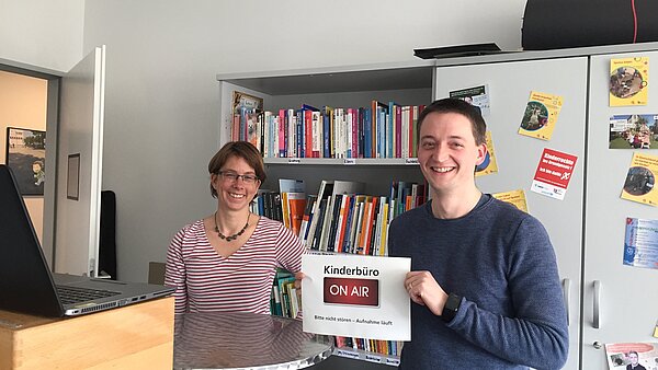 Zwei Personen in einem Büro zeigen ein Schild mit der Aufschrift Kinderbüro On Air