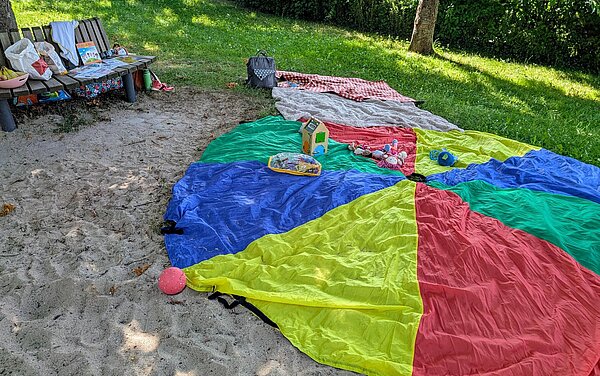 Bunter Teppich auf Sandboden, seitlich Holzbank beides bestückt mit Spielsachen für Kinder.