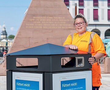 Blick auf eine Mitarbeiterin des Team Sauberes Karlsruhe vor einem Abfallbehälter. Im Hintergrund die Pyramide auf dem Marktplatz.