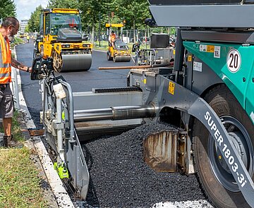 Bei der Baustelle an der Ludwig-Erhard-Allee wird zur Fahrbahnsanierung ein neuartiger Niedertemperaturasphalt auf der Fahrbahn aufgebracht.