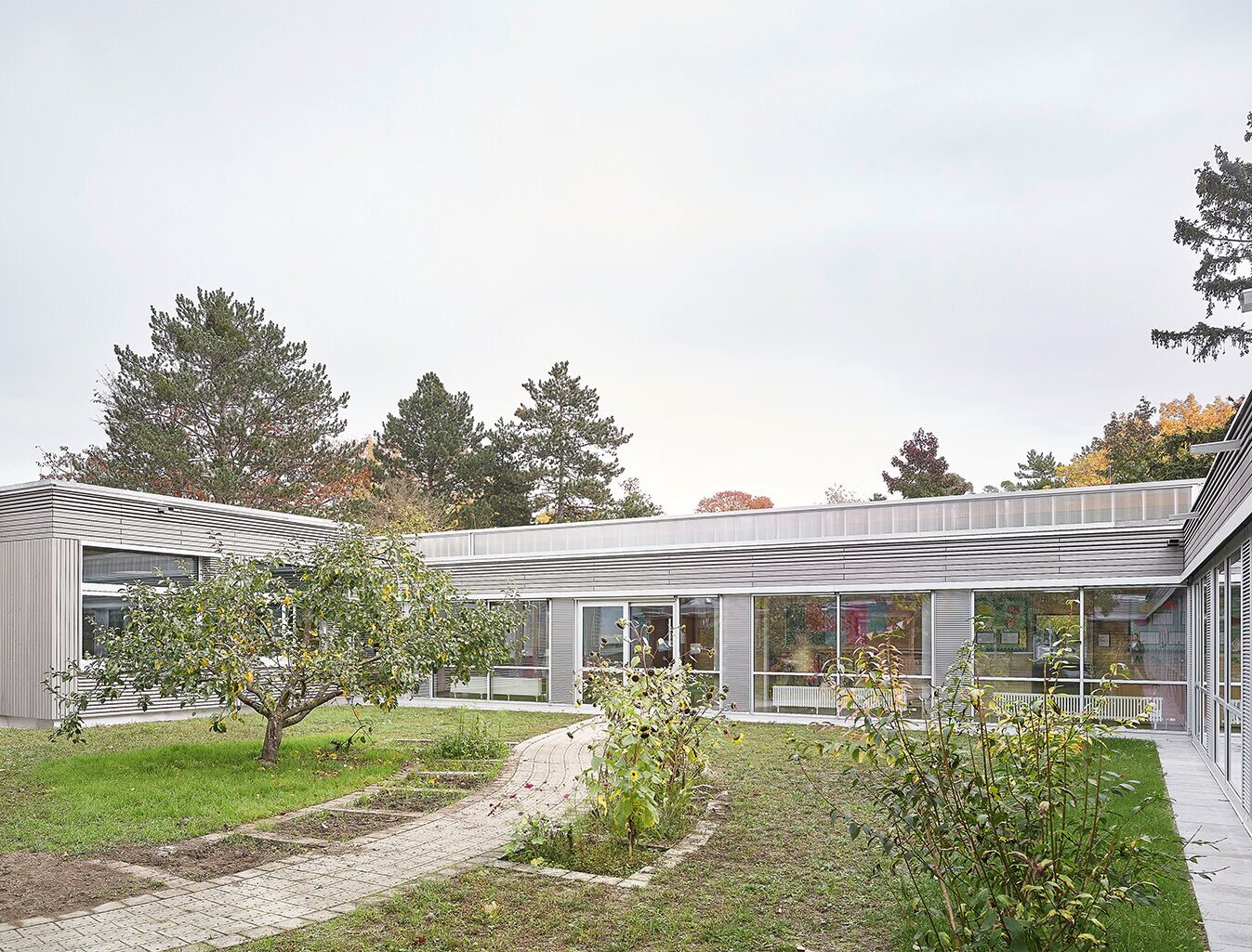 Das Bild zeigt die grau lasierte Nordfassade aus Holz mit großen Fensterflächen und den Schulgarten.