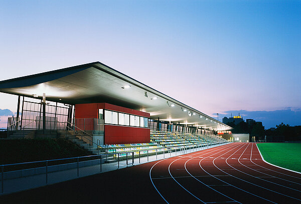 Zu sehen ist die beleuchtete Tribüne mit Wettkampfbüro bei Dämmerung sowie ein Teil der Tartanbahn.