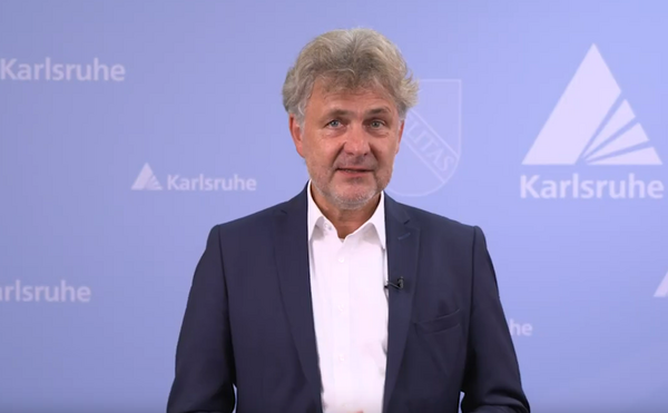 Oberbürgermeister Dr. Frank Mentrup vor blauem Hintergrund mit Fächerwappen der Stadt.