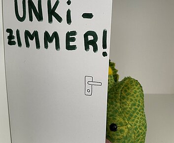 Chamäleon versteckt sich hinter Papptür mit Aufschrift Unkizimmer