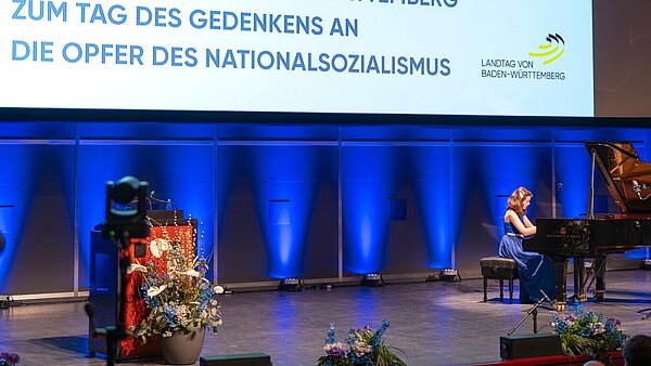 Leinwand im Konzerthaus mit Überschrift "Gedenkstunde des Landtags von Baden-Württemberg zum Tag des Gedenken an die Opfer des Nationalsozialismus". Auf der Bühne spielt Fenella Bockmaier Klavier.