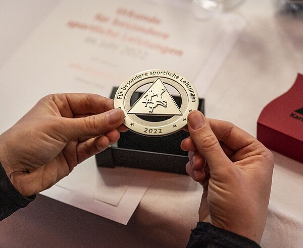 Zwei Hände halten eine runde, silberne Medaille.
