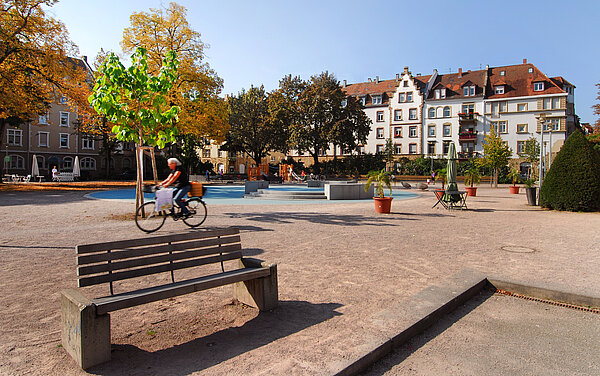 Das Bild zeigt den Fliederplatz im Stadtteil Mühlburg mit einer Bank im Vordergrund und Wohnhäuser im Hintergrund. 