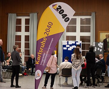 Im Vordergrund ist eine Dropflag mit Logo von Karlsruhe 2040 zu sehen. Im Hintergrund ist eine Menschengruppe an Stellwänden, vertieft in Diskussionen zu erkennen.