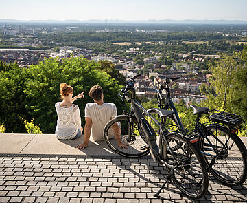 Zwei Personen sitzen auf der Turmbergterrasse. Neben ihnen stehen zwei Fahrräder. Sie blicken auf das Stadtgebiet Karlsruhed
