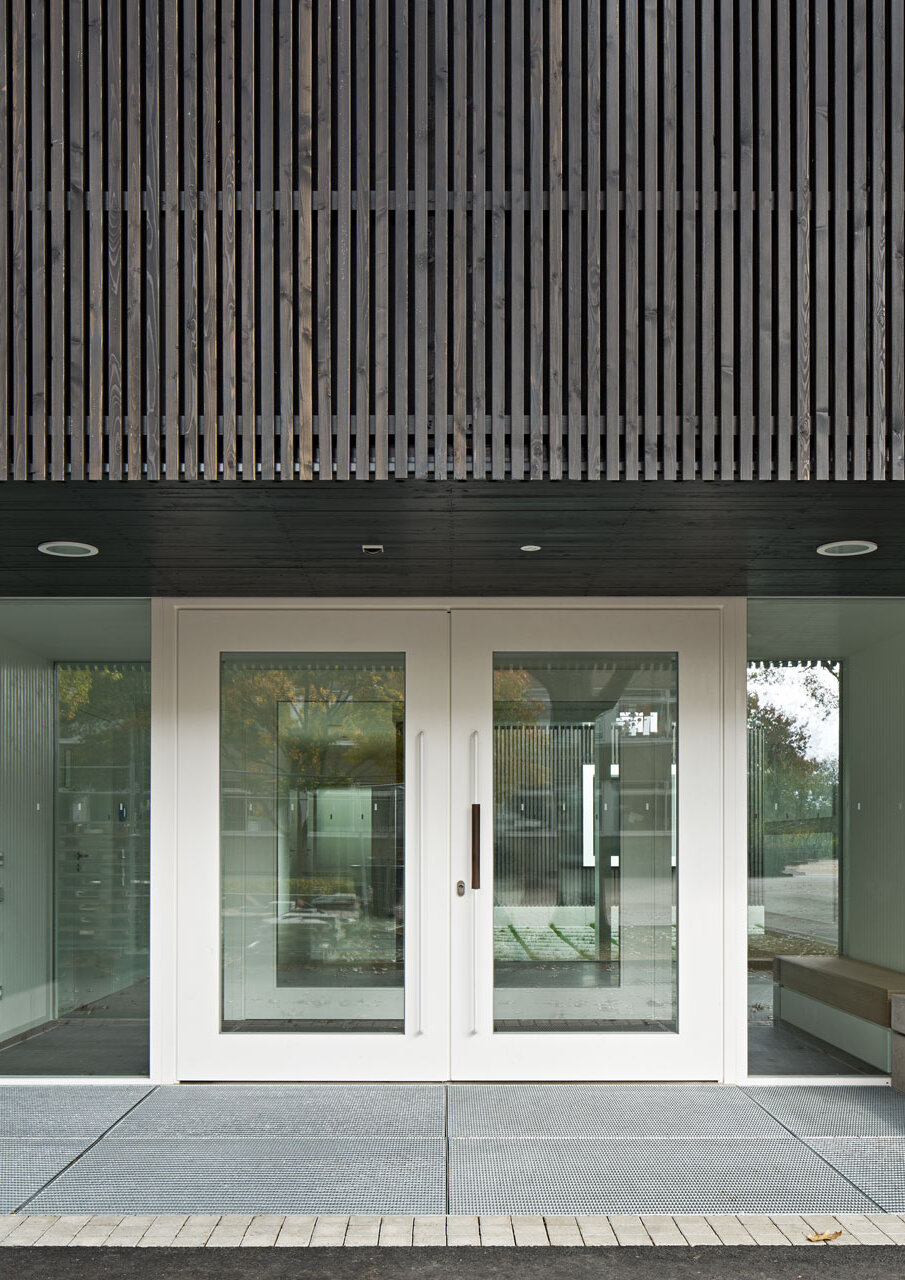 Nahaufnahme des Eingangsbereichs mit Glastüren im weißen Holzrahmen und umliegend ein Teil der dunklen Holzlattenfassade.