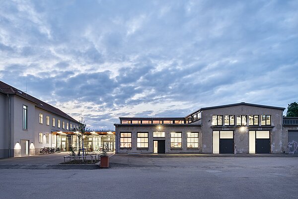 Das Bild zeigt die beleuchtete Südostfassade des Zwischenbaus und ein Teil der ehemaligen Reparaturhalle bei Dämmerung. 