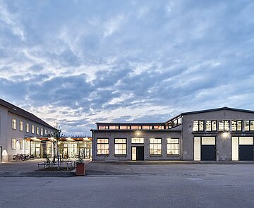 Das Bild zeigt die beleuchtete Südostfassade des Zwischenbaus und ein Teil der ehemaligen Reparaturhalle bei Dämmerung. 