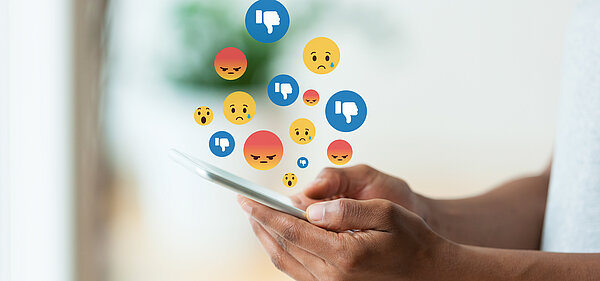 Person hält Handy in den Händen über dem traurige und wütende Emojis zu sehen sind. 