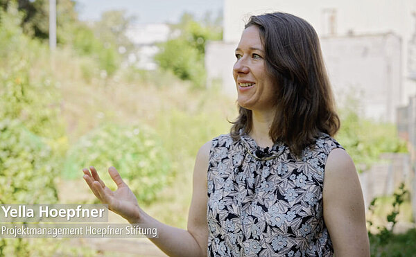 Videostill zeigt Yella Hoepfner, Projektmanagerin der Hoepfner-Stiftung mit der alten Malzfabrik im Hintergrund.