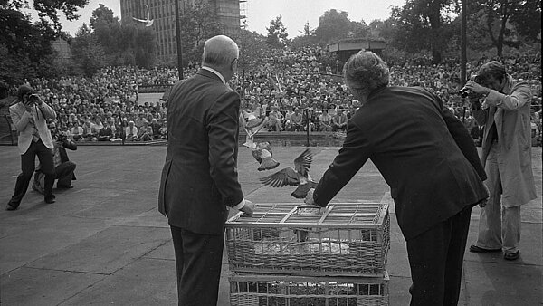 Der Oberbürgermeister von Nancy Marcel Martin (links) und Oberbürgermeister Otto Dullenkopf (rechts) auf der Seebühne im Stadtgarten bei der Aussendung von Brieftauben. Im Hintergrund dichtes Publikum.