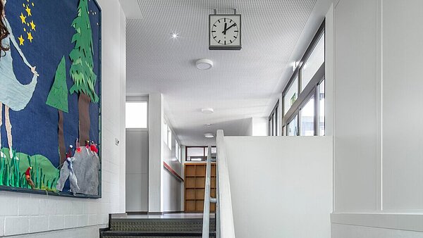 Blick vom Treppenpodest die Treppe hinauf in den hellen Flur des Erdgeschosses mit neuer Akustikdecke und energieeffizienten LED-Leuchten.