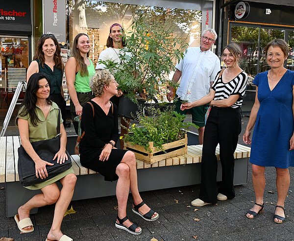Acht Personen sitzen auf einer Holzbank im Passagehof. Zwischen ihnen ist eine Kiste mit Staudenpflanzen.