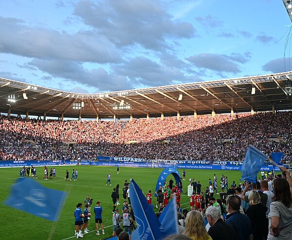 Ansicht des Spielfelds und dem Publikum das blauhe Fähnchen schwenkt.