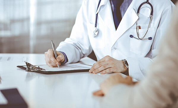 Unbekannter männlicher Arzt und Patientin diskutieren etwas, während sie in einer Klinik sitzen und einen Klemmbrett verwenden.