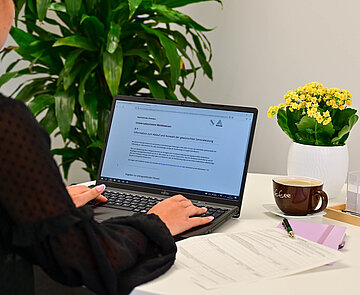 Das Bild zeigt eine Person, die an einem Schreibtisch sitzt. Sie hat vor sich ein Notebook, daneben liegen Formulare.