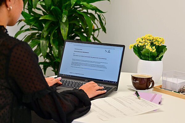 Das Bild zeigt eine Person, die an einem Schreibtisch sitzt. Sie hat vor sich ein Notebook, daneben liegen Formulare.